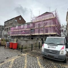 A building under renovation is enveloped in pink commercial scaffolding. Nearby, a white work van with "A Star Access" rests on the cobblestone street. Overcast skies loom above, while a red trash bin sits close to the busy construction site.