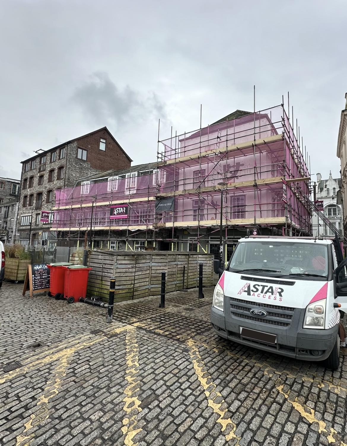 A building under renovation is enveloped in pink commercial scaffolding. Nearby, a white work van with "A Star Access" rests on the cobblestone street. Overcast skies loom above, while a red trash bin sits close to the busy construction site.