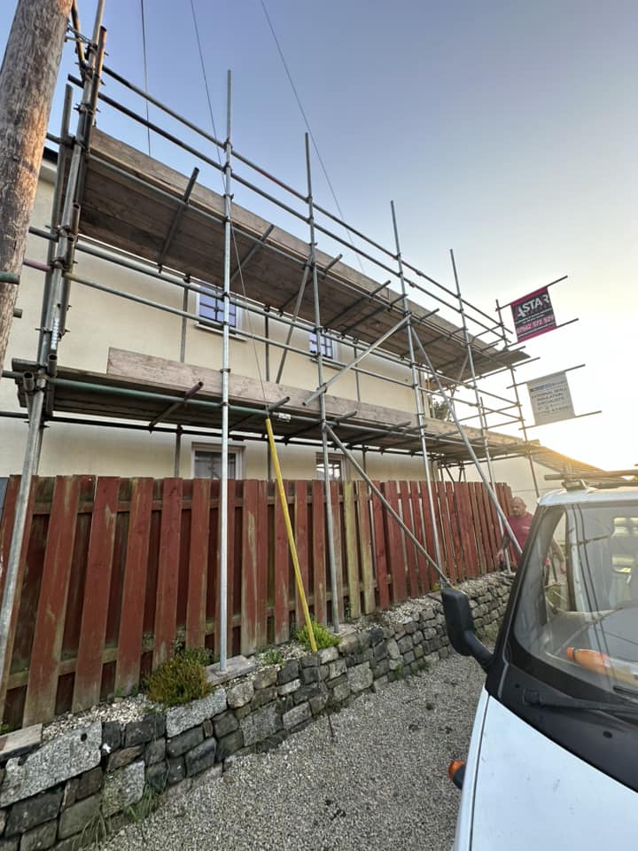A building under construction features commercial scaffolding in place. A wooden fence stands in the foreground, while a parked vehicle is partially visible on the right. The clear sky, bathed in sunlight, suggests it's early morning or late afternoon.