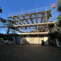 A large metal commercial scaffolding structure is set up in front of a building. A clear blue sky stretches above, and trees flank either side. A ladder leans against the building, while a flag with text proudly waves atop the scaffolding.
