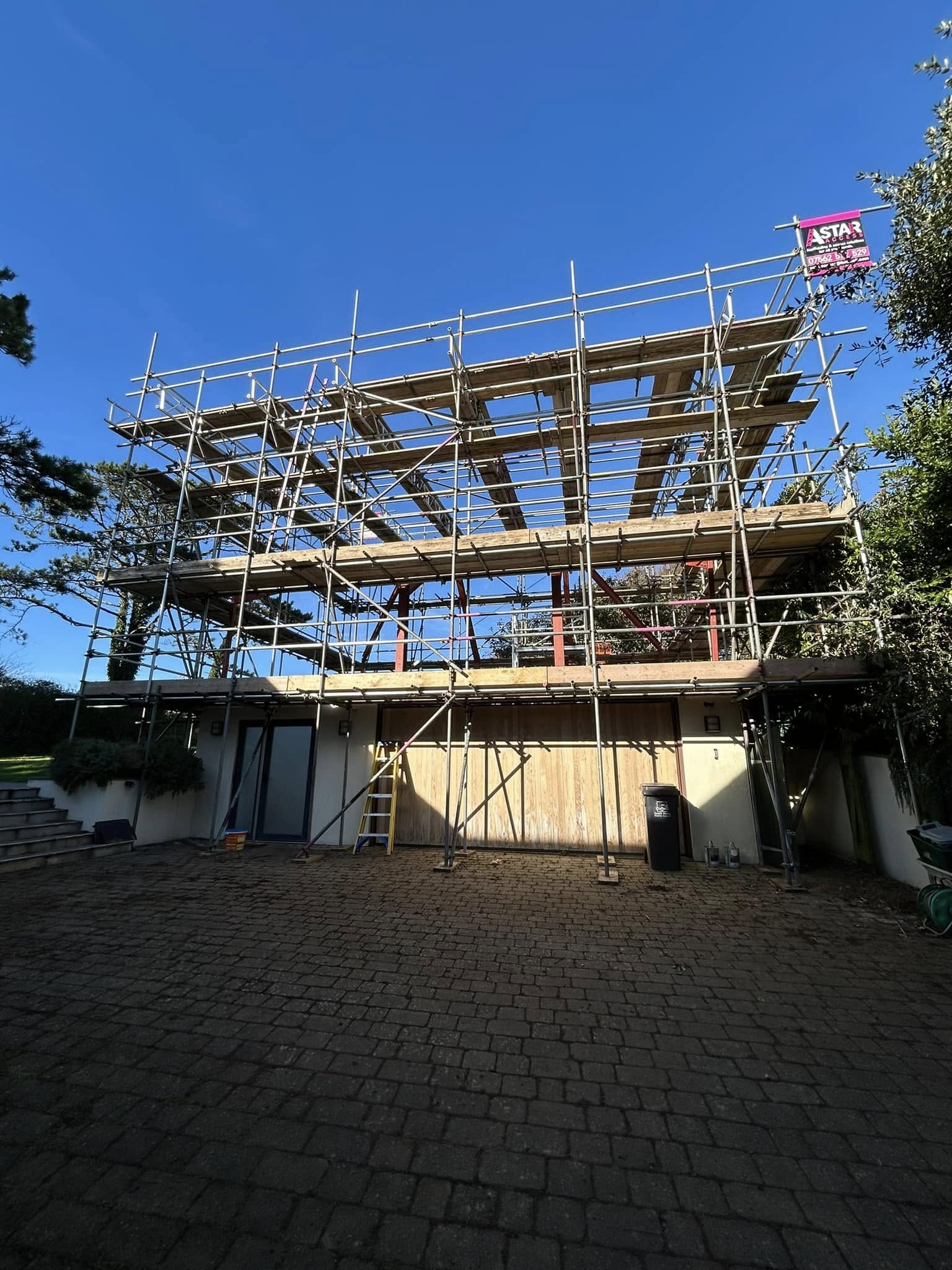 A large metal commercial scaffolding structure is set up in front of a building. A clear blue sky stretches above, and trees flank either side. A ladder leans against the building, while a flag with text proudly waves atop the scaffolding.