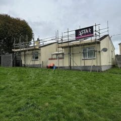 A small beige house with domestic scaffolding on the roof sits on a grassy lawn. A sign reading "AStar" is displayed. A satellite dish and some construction materials are visible near the building under the cloudy sky.